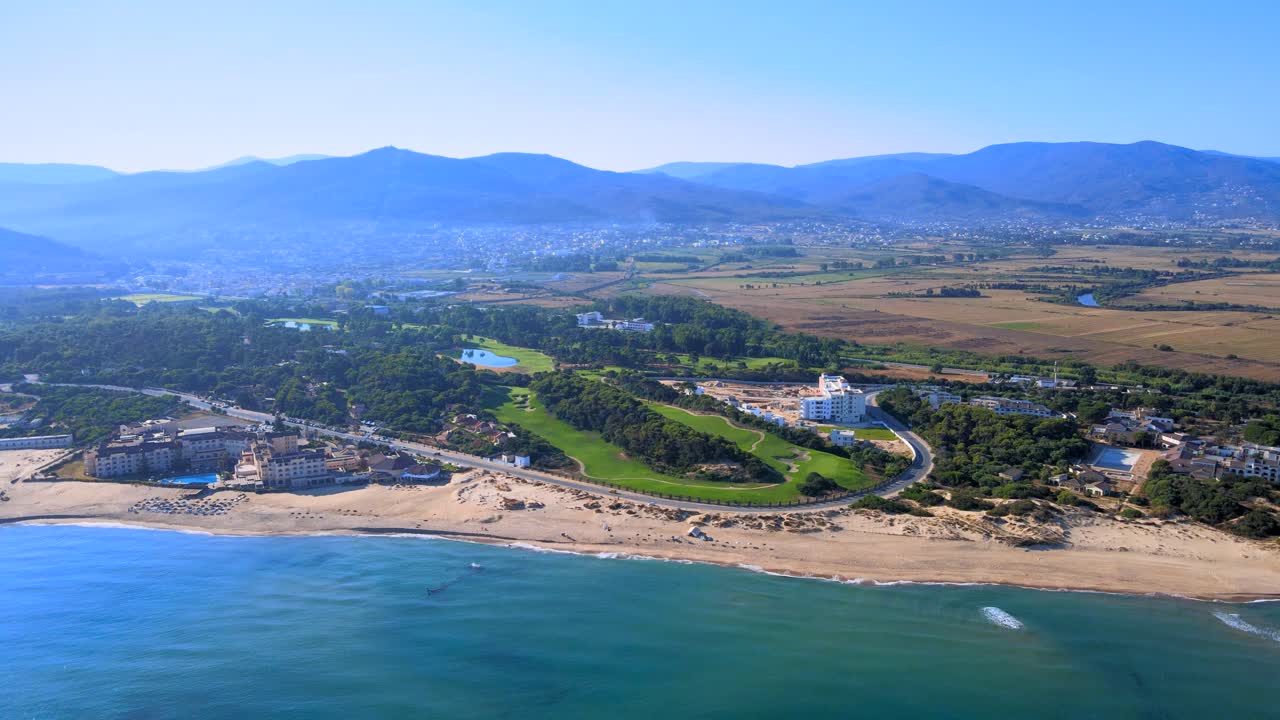 The image depicts a breathtaking scenic view of fields on the beach with mountains towering above them. A city is visible below while a river flows through it.