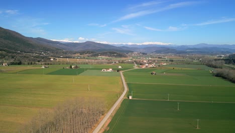 Drone-aerial-footage-of-a-narrow-road-with-cultivated-green-fields-on-the-sides-and-snow-capped-mountains-in-the-background,-Crane-plane-pointing-up