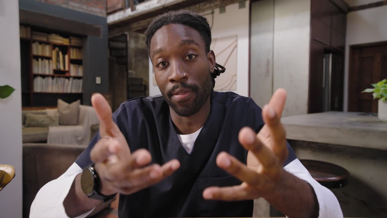 African male in scrubs leaning in, adjusting framing, settling and explaining on camera at table