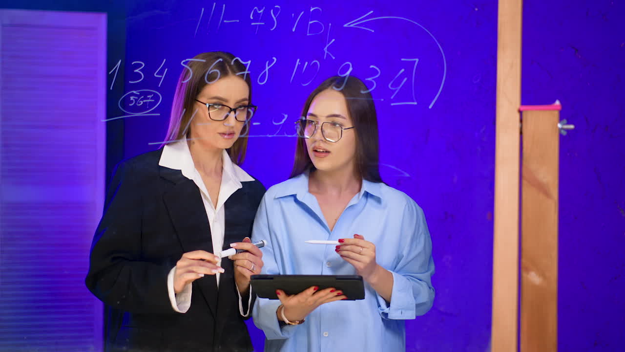Caucasian ladies look at tablet, communicate and compare the numbers on the glass board. Woman in black jacket writes on the board. Business concept.
