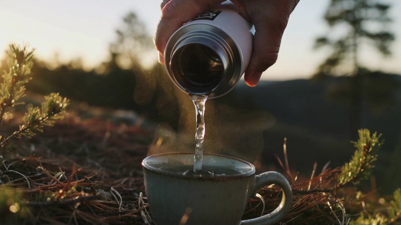 Thermos metallico inclinato, una mano versa del liquido per riempire una tazza di ceramica su un terreno di aghi di pino, con vapore che sale.