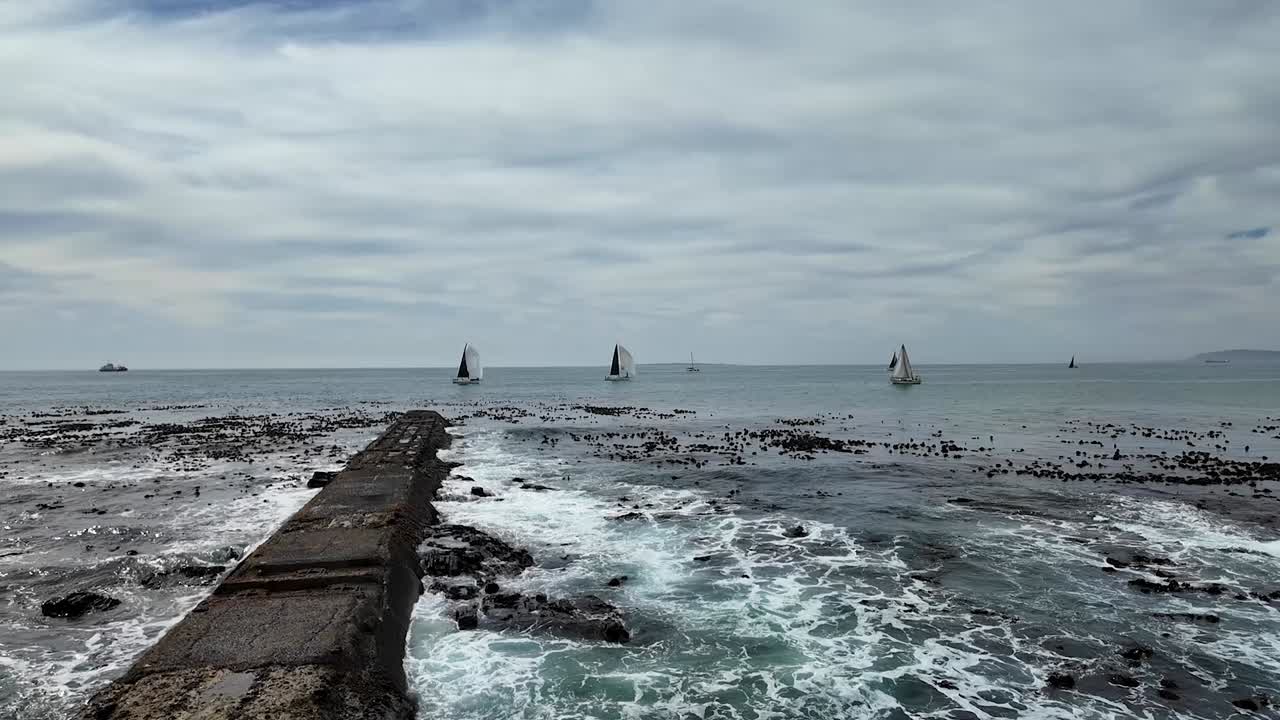 Sailboats on a choppy sea near a stone jetty, Cape Town, South Africa.