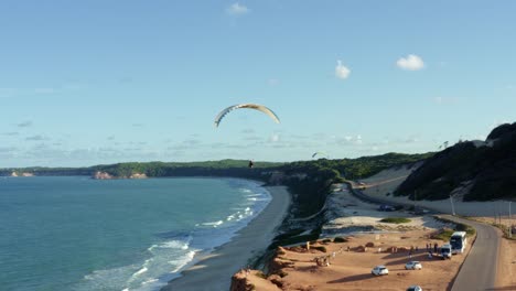 tomada aérea de un parapente volando sobre la impresionante costa tropical del noreste de brasil cerca de pipa en río grande do norte con grandes dunas de arena, acantilados sobre el océano y follaje verde