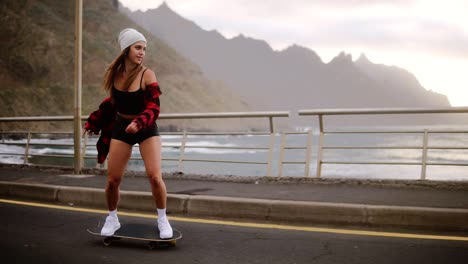 hermosa chica musculosa monta una patineta y cae ligeramente de ella, pero se mantiene en el suelo. costa, océano y colinas en el fondo