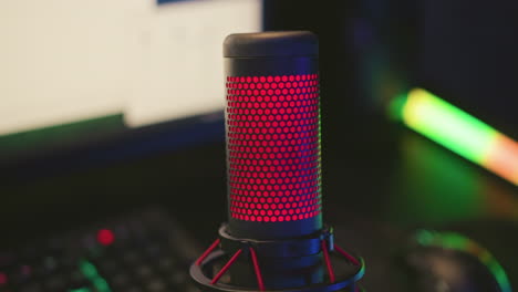 Close up condenser microphone illuminated with red light positioned in front of blurred computer setup including monitor screen, keyboard, mouse, and multicolor LED strip