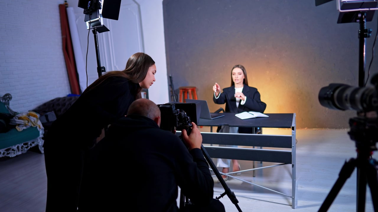 Instruktør og assistent gennemgår optagelser under studievideooptagelse. Produktionshold kigger på kameramonitor, mens de filmer kvindelig vært ved skrivebord.