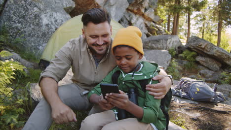 happy father and mixed race son using smartphone at campsite
