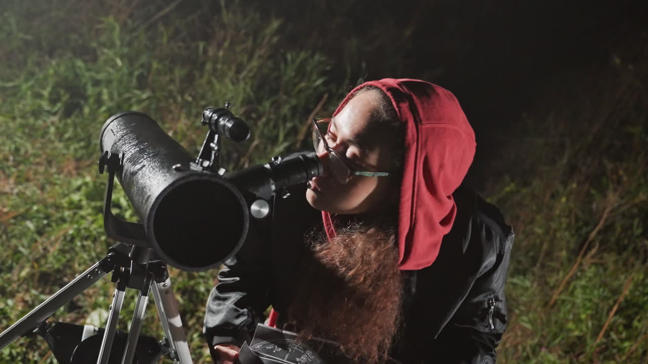 night stargazer with telescope, hooded observer consults star chart beside large reflector on tripod in dewsoaked field bearded face peers through eyepiece, adjusting focus while taking notes