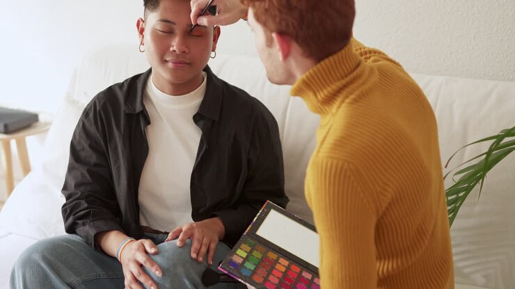 A person laughs joyfully while getting makeup applied by another, featuring a rainbow bracelet