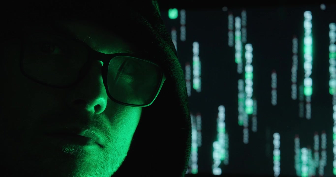Close-up of a hooded man wearing glasses with glowing digital code in the background. Concept of hacking, cybersecurity, anonymity, data protection, and cybercrime.