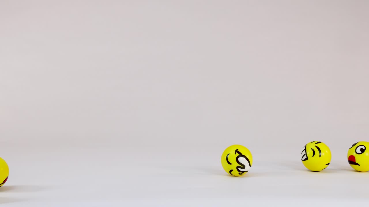 Yellow balls with various emoji faces bounce and roll across a plain white studio background