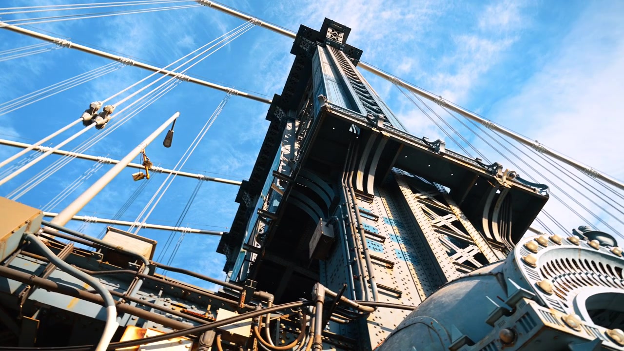 Manhattan Bridge tower and suspension cables from below. Riveted steel beams rise against blue sky streaked with thin clouds.