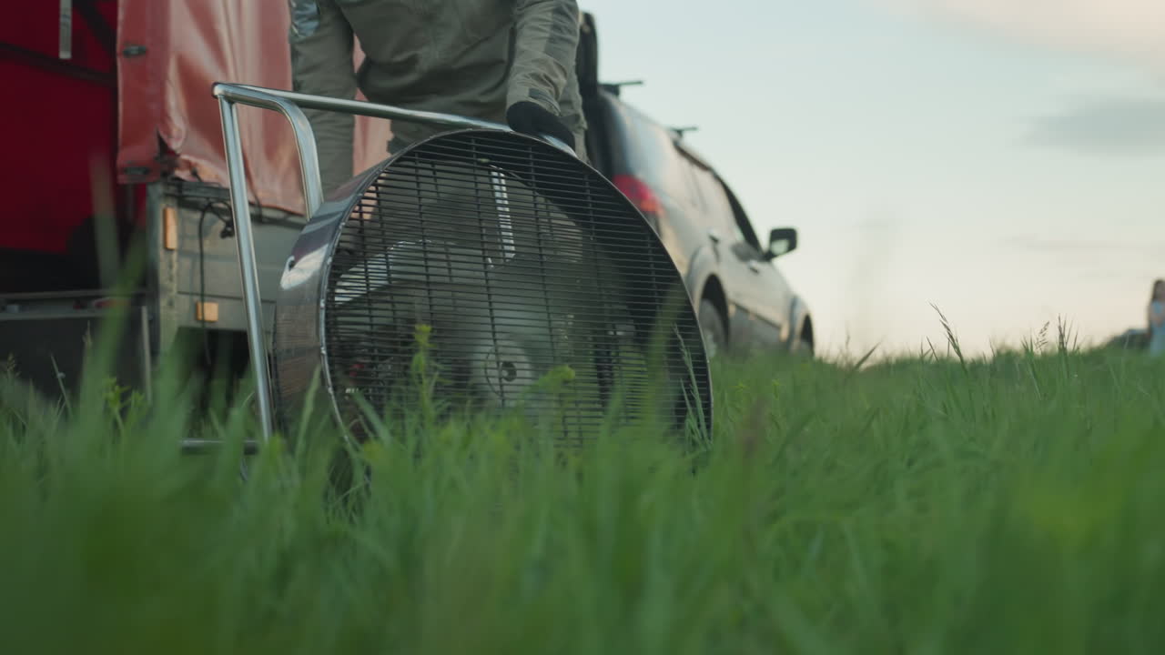 man wearing cap and gloves leans over large fan assembly on grassy field to warm hot air balloon engine before sunrise flight as crew and vehicles stand under cloudy sky over farmland