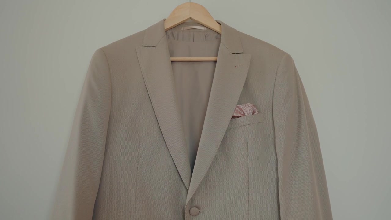 Vista frontale di un'elegante giacca da completo beige su una gruccia di legno con dettaglio di fazzoletto da taschino piegato.