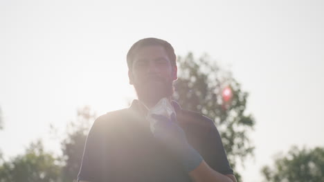 man gardener removing protective mask while sunlight creates lens flare during outdoor work after fumigation in field setting, background trees blurred