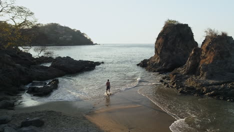 Slowly-rising-aerial-as-man-walks-into-ocean-on-beautiful-secluded-beach,-4K