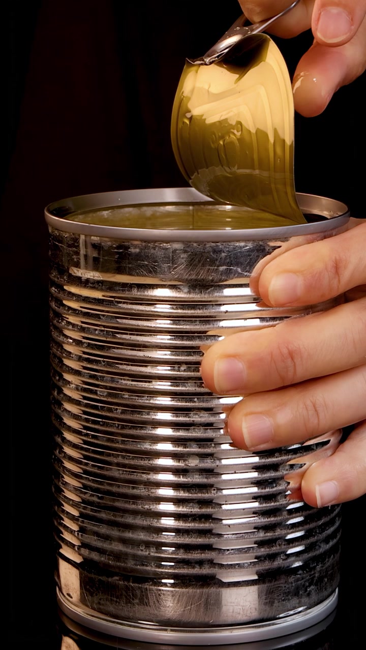 A person’s hands open a metal pull-tab can, revealing contents inside. Studio lighting highlights the process, with a close-up, steady camera and dark backdrop