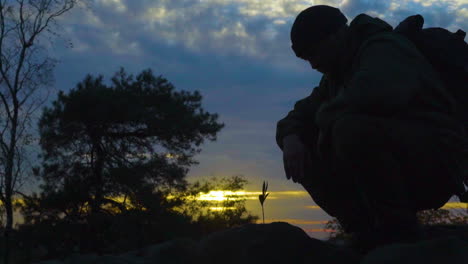 silhouette of man in wilderness looking at growing plant during sunset