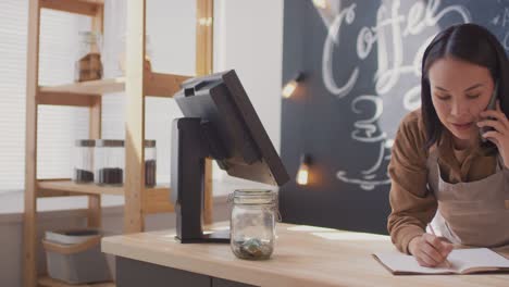 Female-Waitress-Taking-Notes-And-Talking-On-The-Phone-On-The-Counter-In-A-Coffee-Shop