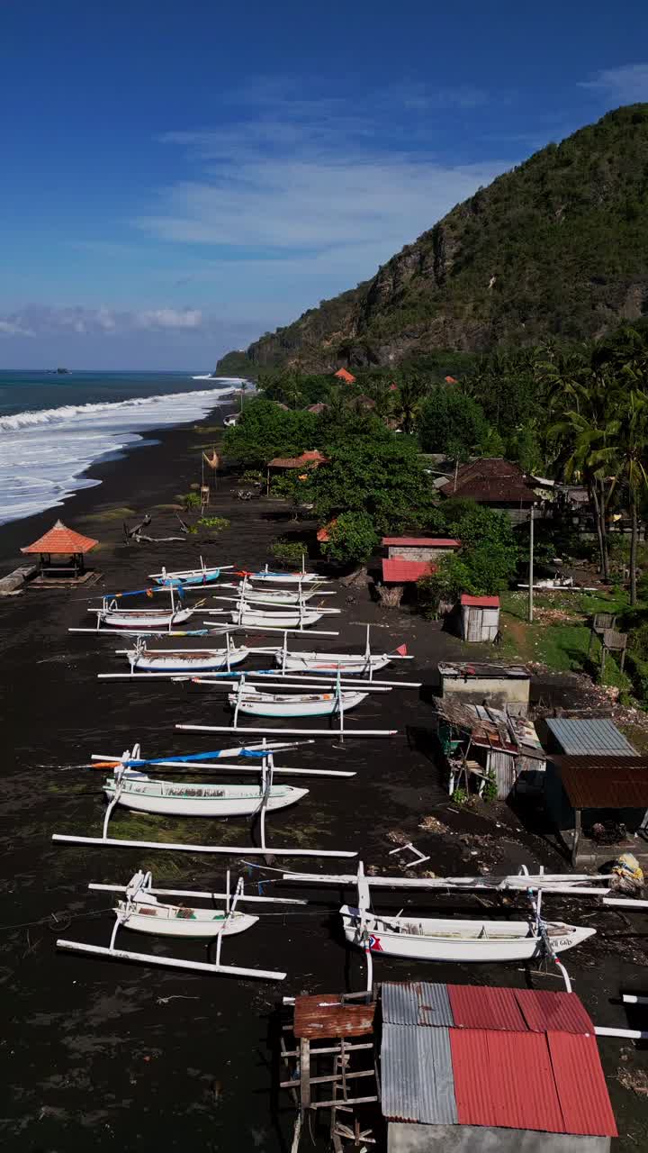 Balinese fishing boats sit along a volcanic sand beach while ocean waves gently break and green hills frame the coastline captured in stunning aerial drone footage revealing the islands natural charm