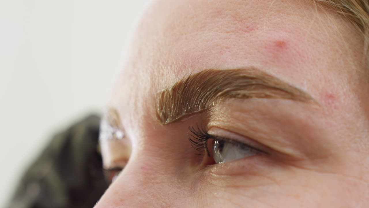 Close up of beautician using cotton swab to clean excess substance from beneath client eyebrow during beauty procedure. Focus on brow texture and precision care in personal grooming session