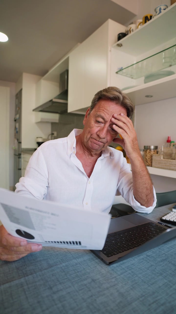 Uomo anziano che esamina documenti finanziari e lavora al laptop a casa.