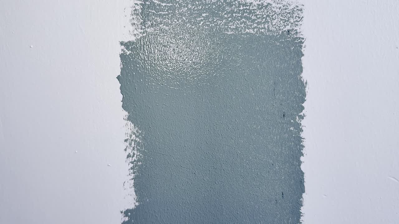 Primo piano di un rullo per pittura che applica uno strato di vernice grigia su una parete bianca liscia, mostrando le pennellate fresche.