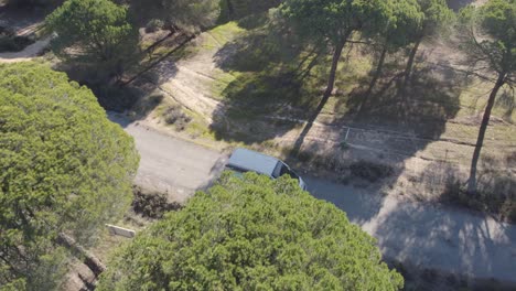 Aerial-view-orbiting-motorhome-campervan-driving-narrow-road-through-woodland-trees-in-rural-countryside-road-trip,-Spain