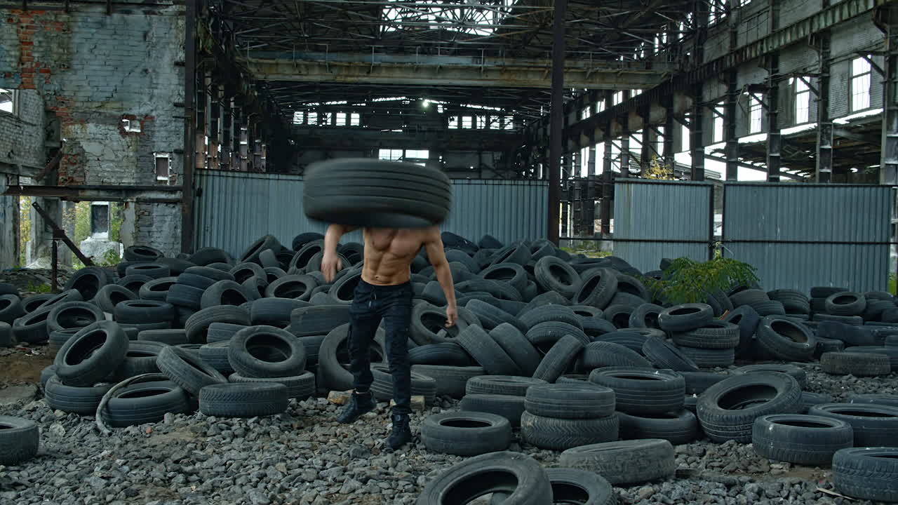 Training session of a sportsman in abandoned place. Strong man without shirt scattering over old car tires during his workout on ruined factory background.