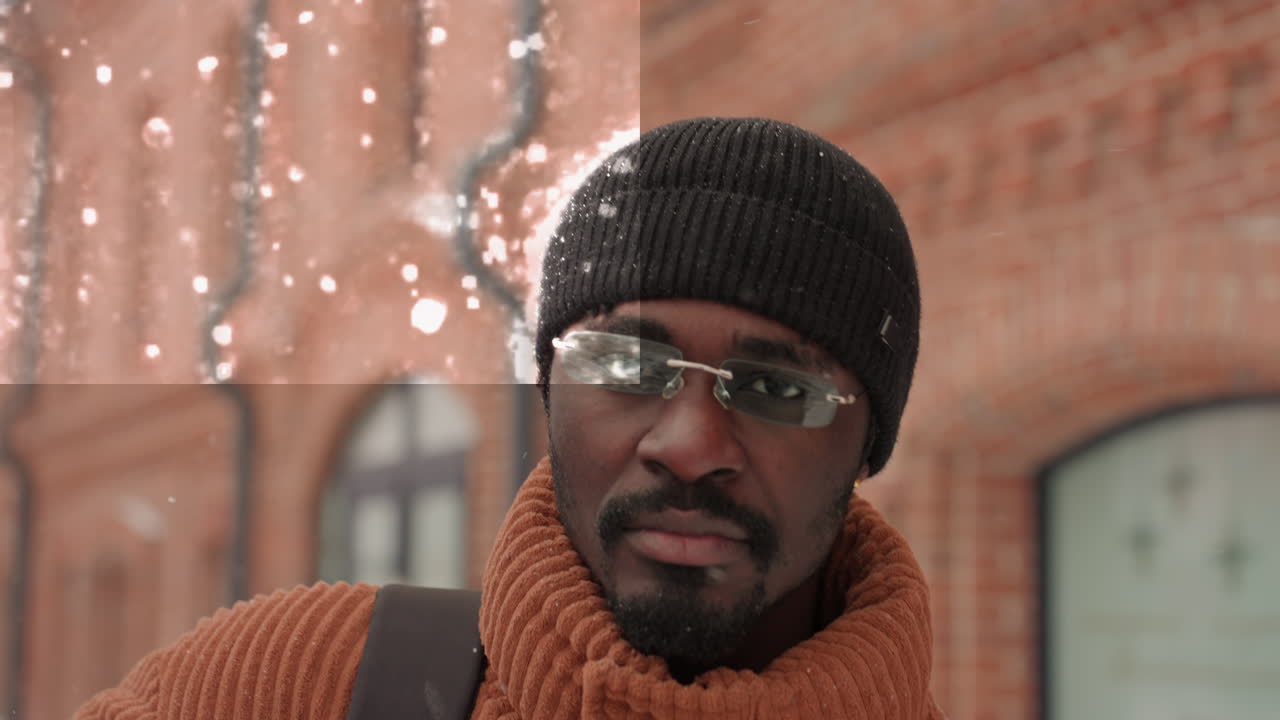 Schwarzer Mann im fallenden Schnee vor roter Ziegelfassade, Strickmütze mit Schneeflocken bestäubt, Brille reflektiert Winterhimmel, orangefarbener Pullover und Taschenriemen sichtbar, gelassener Ausdruck, filmisches Bokeh