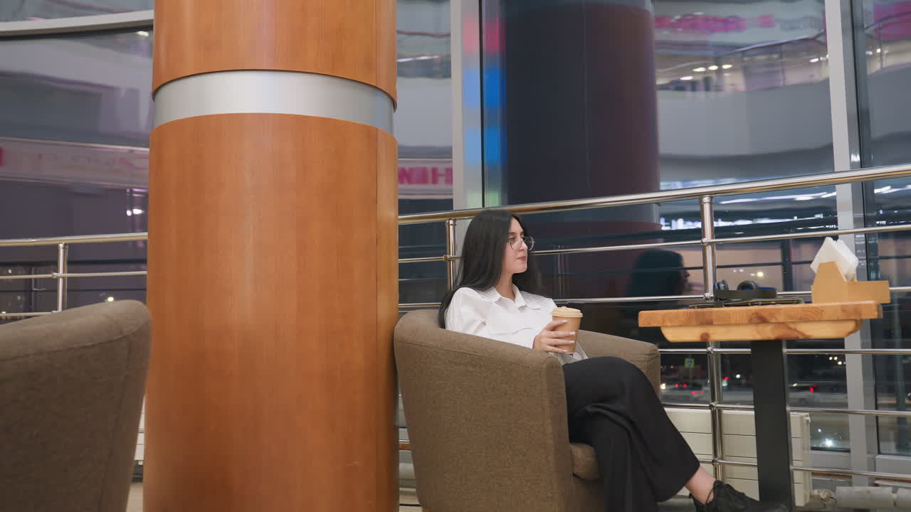 investor sips drink and lowers cup while staring thoughtfully through glass panel with night traffic visible outside, seated in cozy lounge space with tablet and headset placed on wooden table