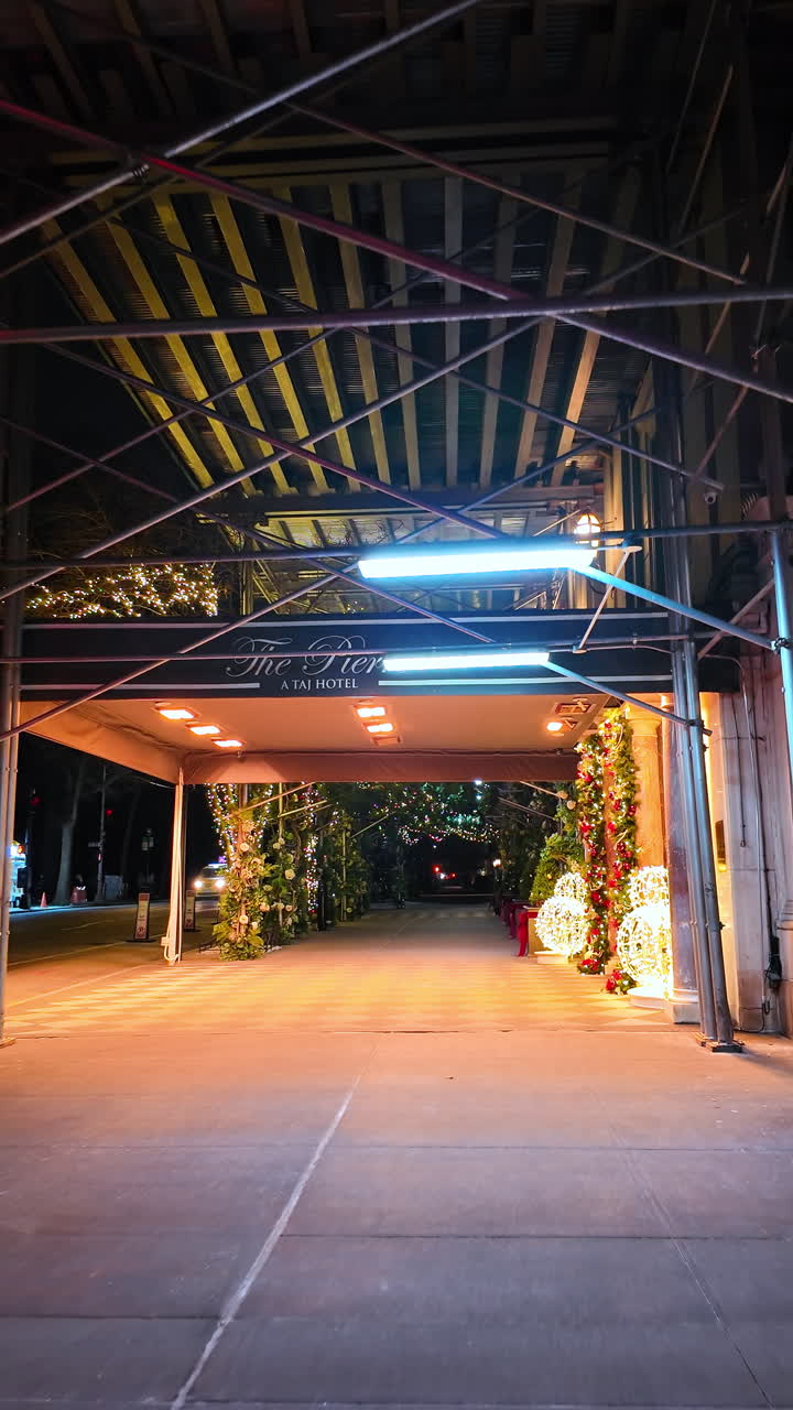 Nueva York, EE. UU., 25 de diciembre de 2025: Acera bajo andamios de construcción con decoraciones navideñas. Vista en perspectiva de un pasillo de acera iluminado cubierto por andamios metálicos cerca de la entrada de un hotel de lujo por la noche.