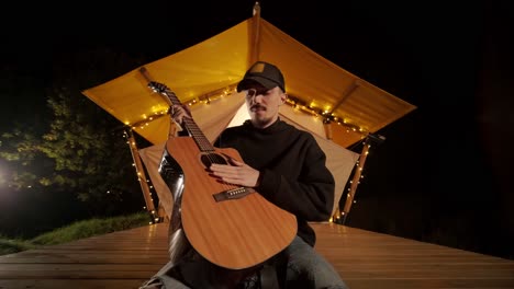 man plays the guitar and sings songs in a tent with light bulbs