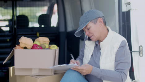 delivery woman talking with customer on phone in a van and writing on clipboard next to grocery box