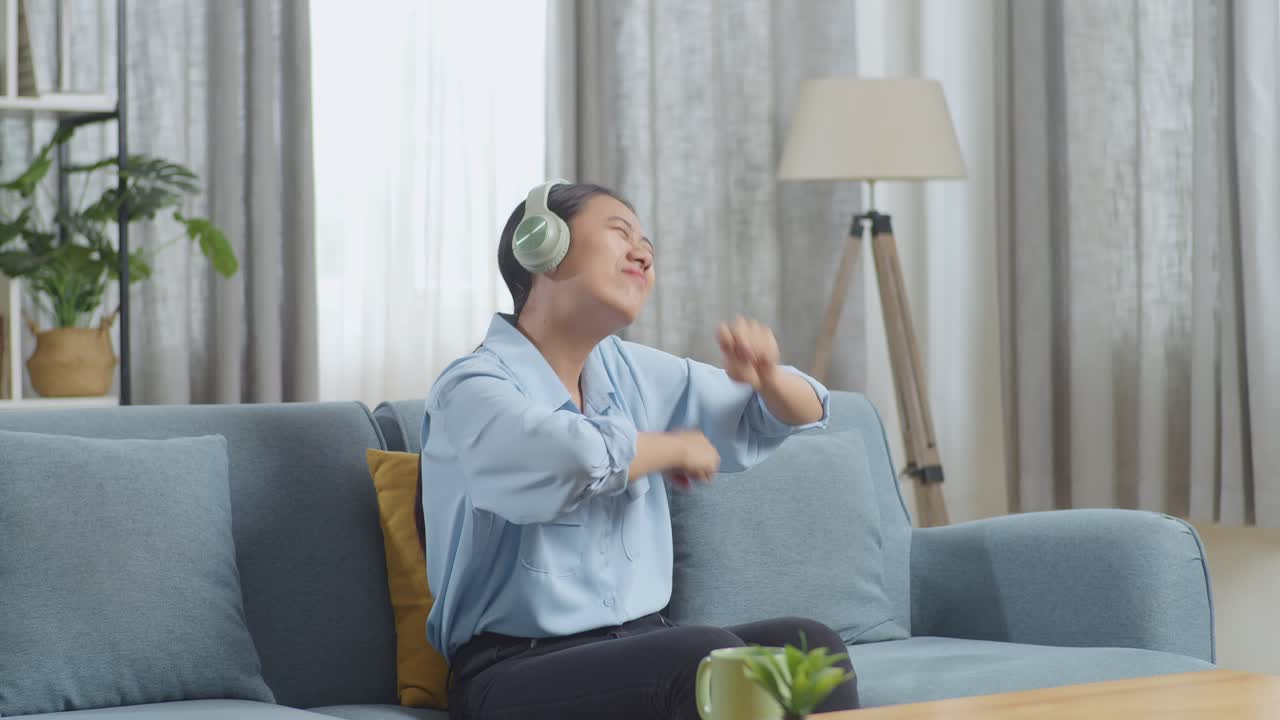 헤드폰 을 착용 한 아시아인 여자 가 거실 의 소파 에 앉아 음악 을 듣고 춤 을 추고 있다