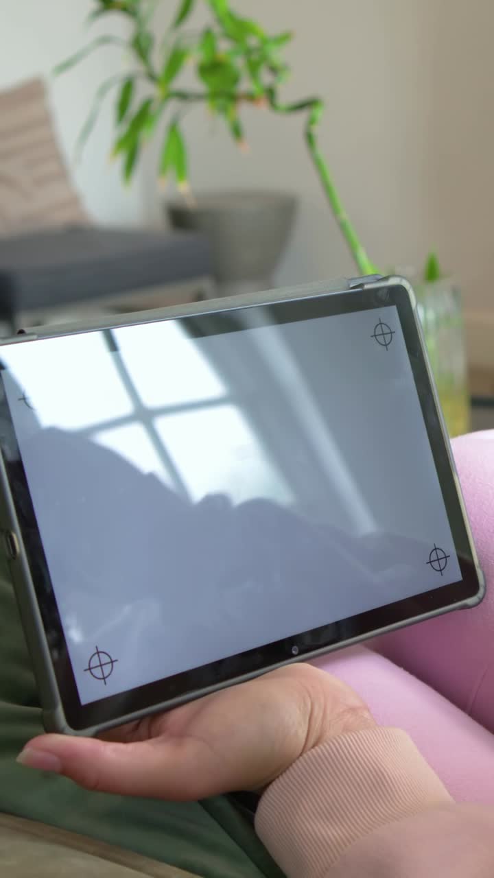 Vídeo vertical: Mulher de rosa com reflexo da janela, inclinando tablet no colo em casa, mostrando mira