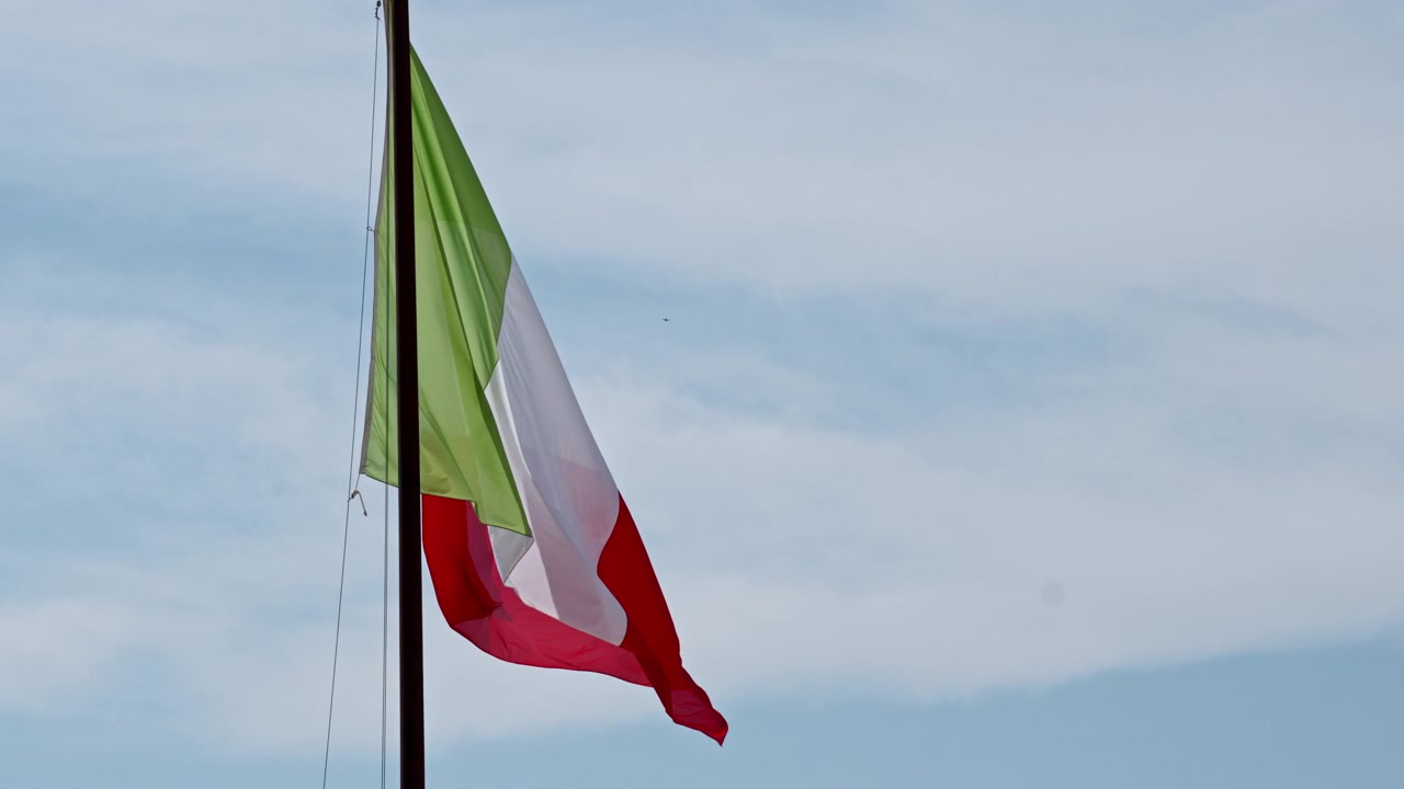 Bandiera italiana verde, bianca e rossa che sventola su un'alta asta con una leggera brezza contro un cielo diurno pallido