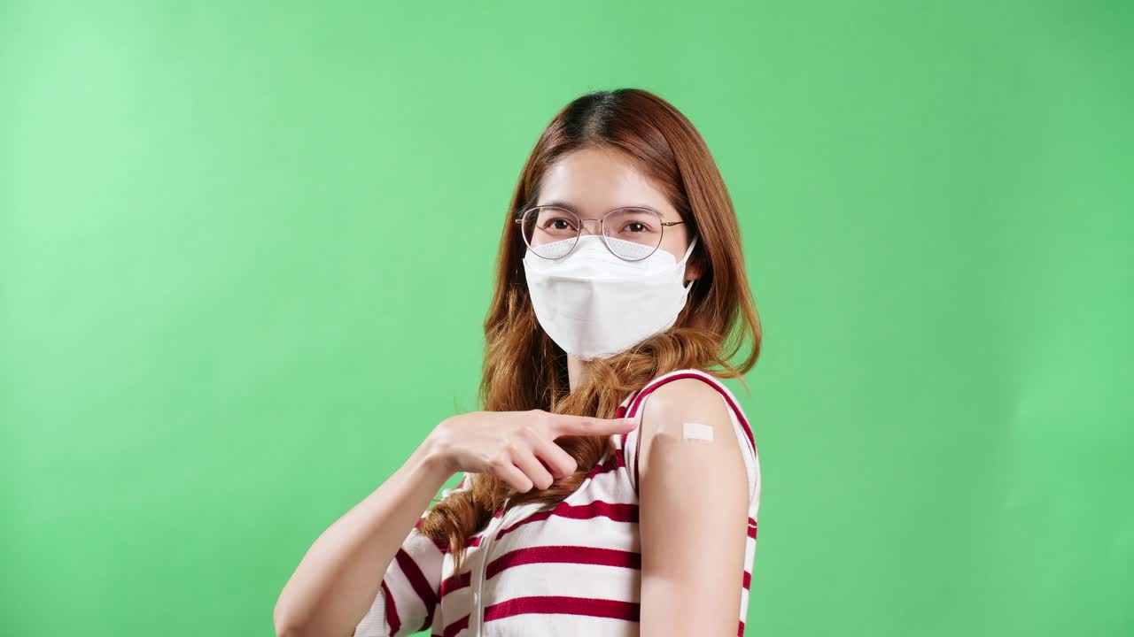 Potret seorang wanita Asia berkacamata tersenyum di balik masker, menunjukkan emosi positif dan ceria setelah divaksinasi, memperlihatkan lengan dengan plester medis di bahunya.