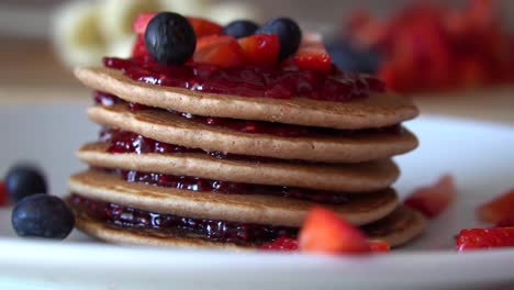 blueberries falling down on top of stacked pancakes with red jam and fresh strawberries, slow motion