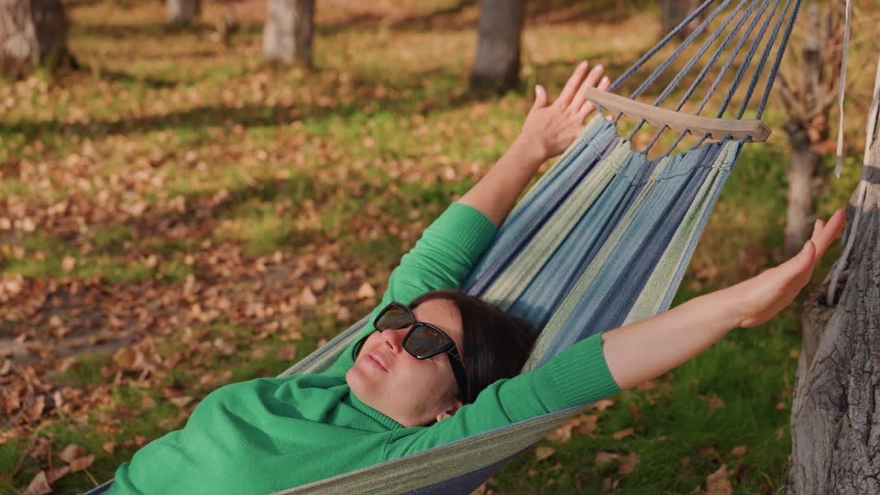 Stretching mattutino sull'amaca da campeggio, uomo che allunga le braccia sopra la testa svegliandosi lentamente in un boschetto illuminato dal sole, indossando occhiali da sole, dondolio delicato contro l'albero, le inquadrature mostrano un allungamento progressivo, sorriso rilassato, movimento sottile.