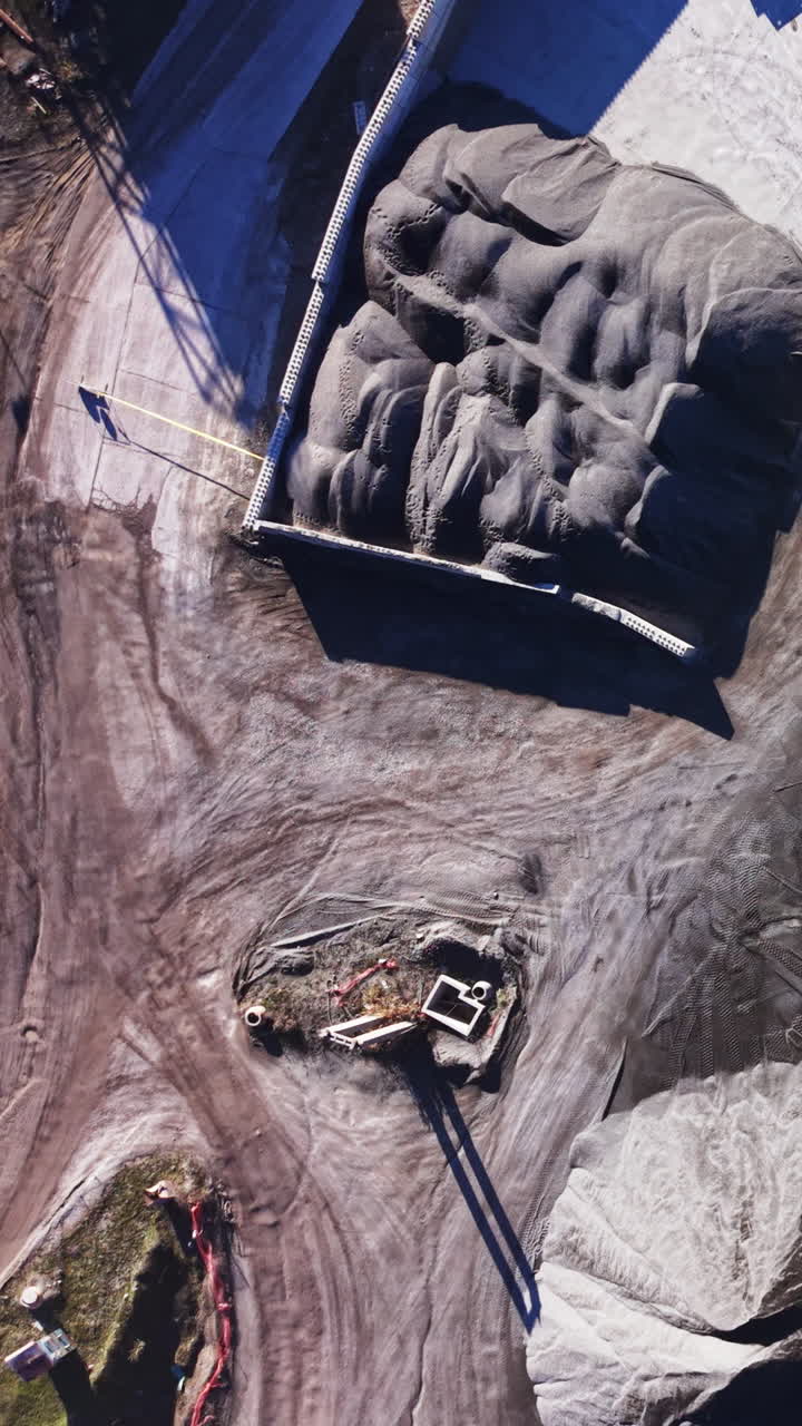 Concrete walled storage site containing massive piles of fine grey sand. Aerial shot of large sand storage area with trucks. Vertical video