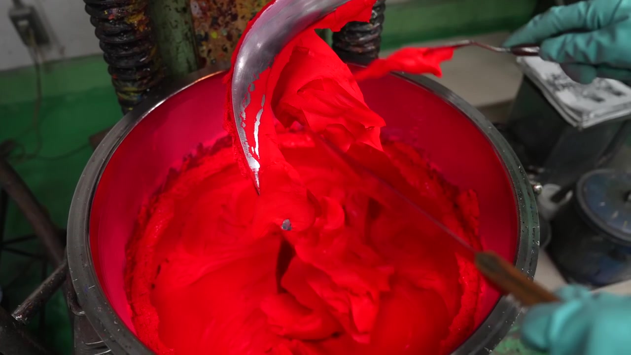 Heavy mixer blade churning bright red oil paint in a stainless bowl while a worker watches beside the machine