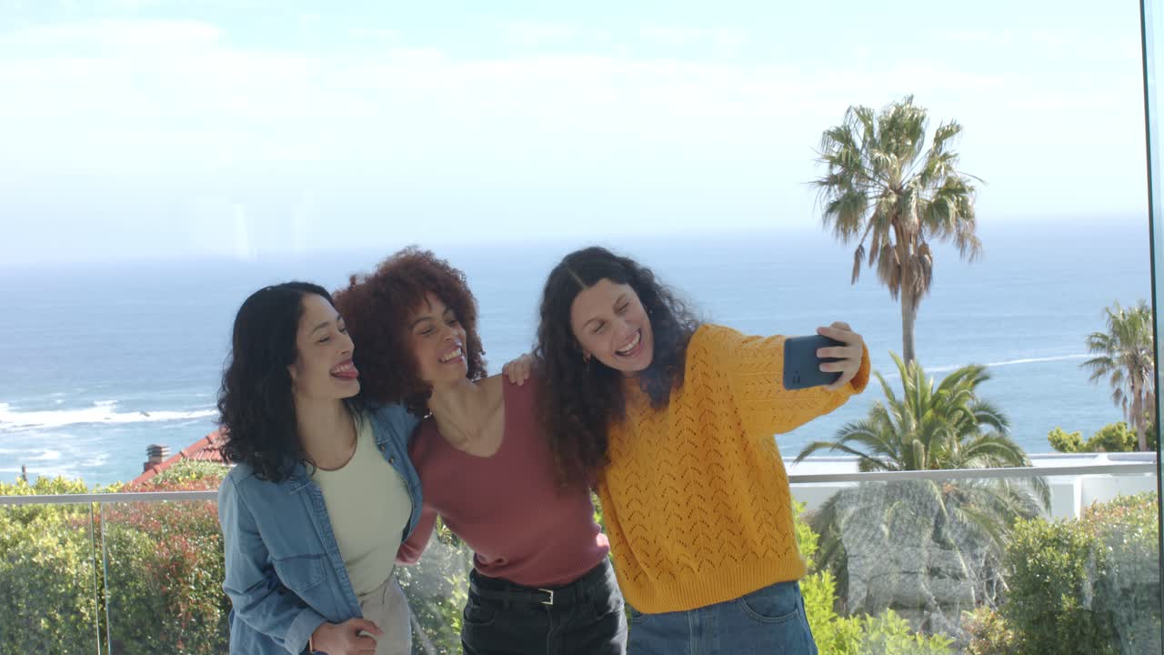 Amiche di diverse etnie che tengono uno smartphone, facendo smorfie per uno scatto selfie su un balcone con ringhiera in vetro.