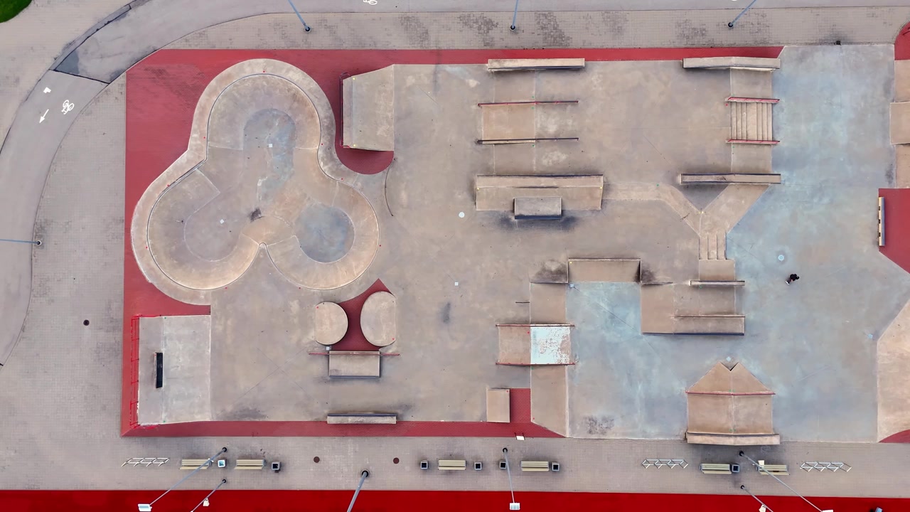 Straight-down drone shot of an empty concrete skatepark with a sculpted bowl, ramps, ledges, stairs and technical obstacles framed by red and blue paving.