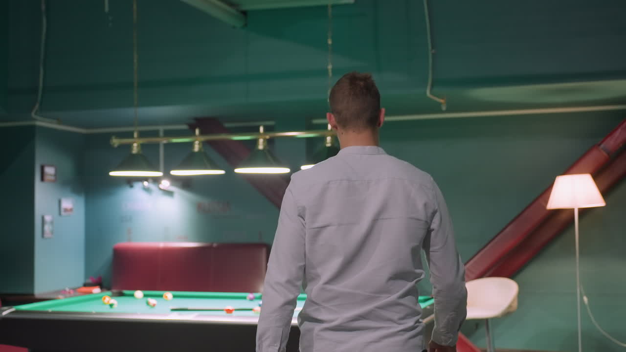 fotografía de cerca de un jugador de billar en camisa blanca entrando en una sala de billar con un interior moderno, acercándose a la mesa de billar y recogiendo el palo de la pilla. mesas de billar verdes, configuración de la lámpara aérea