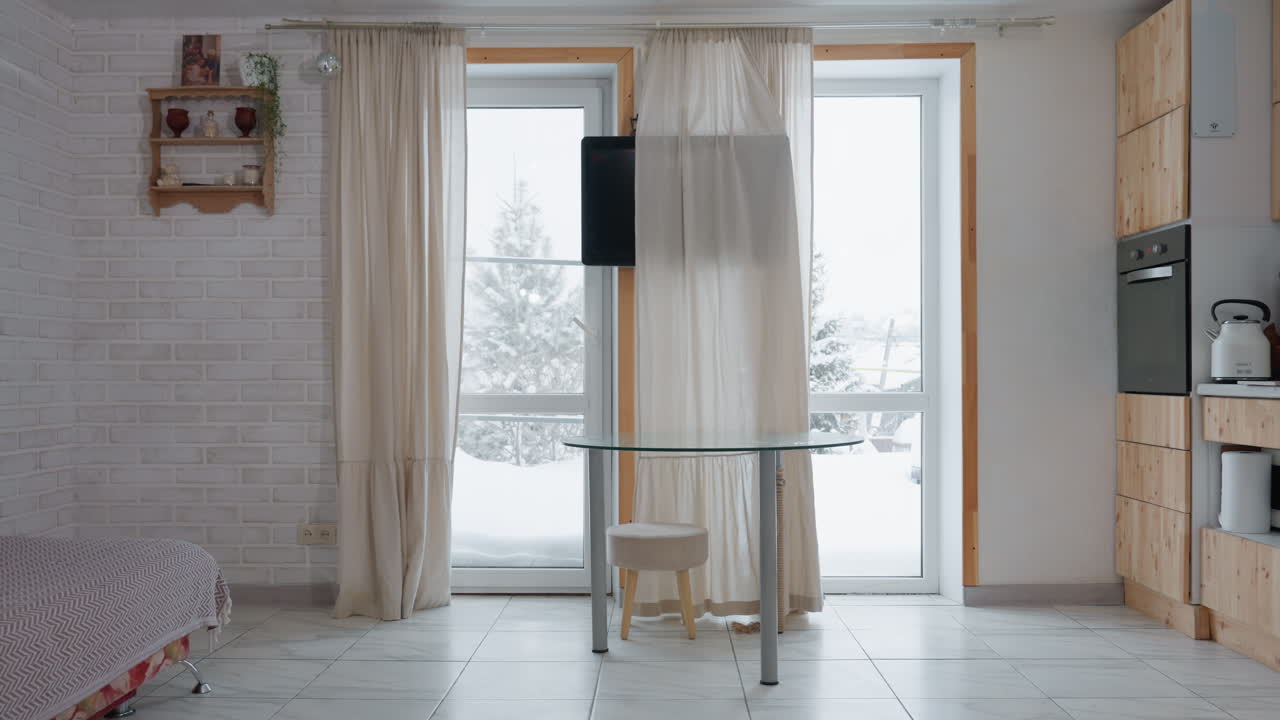 Room with flowing curtains framing transparent window view of snowy landscape, wooden kitchen cabinet, wall mounted television, round glass table with stool, patterned sofa with cushions