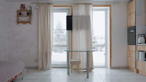 room with flowing curtains framing transparent window view of snowy landscape, wooden kitchen cabinet, wall mounted television, round glass table with stool, patterned sofa with cushions