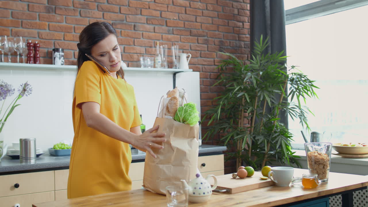 mujer llevando bolsas de comestibles a la cocina, morena terminando una llamada telefónica en casa.