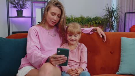 Child-kid-daughter-having-fun-with-mother-or-nanny-enjoying-using-smartphone-watch-cartoons-at-home