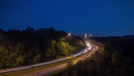 a timelapse long exposure of headlight and tail light trails from vehicle traffic on curved highway road leading towards the downtown toronto skyline at night with trees lining the sides of the road.